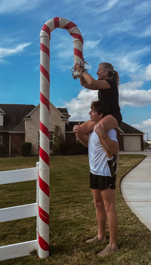 A picture of Eric holding his Mom, Wendy, on his shoulders next to a large red-and-white candy cane. Wendy is holding a string of lights to be wrapped around the candy cane.

