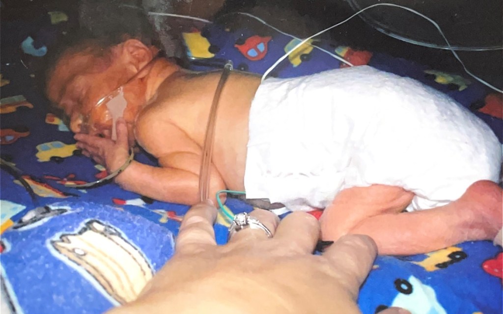 A picture of newborn Eric. He is laying on his stomach on a blue blanket in an incubator. The preemie diaper Eric is wearing is much too large for him.   
