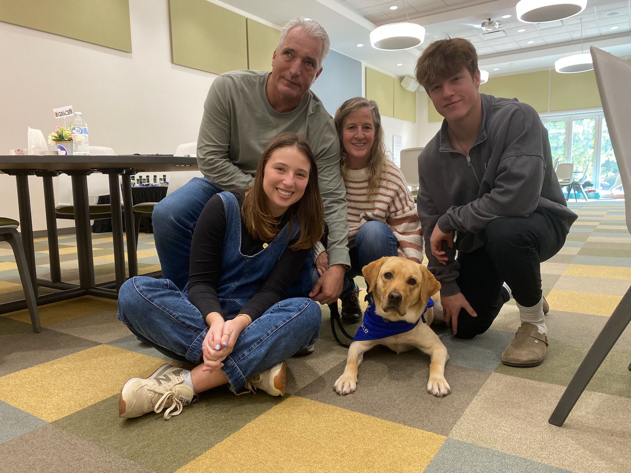 The Royal Family: From left -to-right: Taylor, Mike, Wendy and Eric, kneels next to Southeastern Guide Dog, Grace. Grace is a yellow lab wearing a blue bandana with her name on it. 