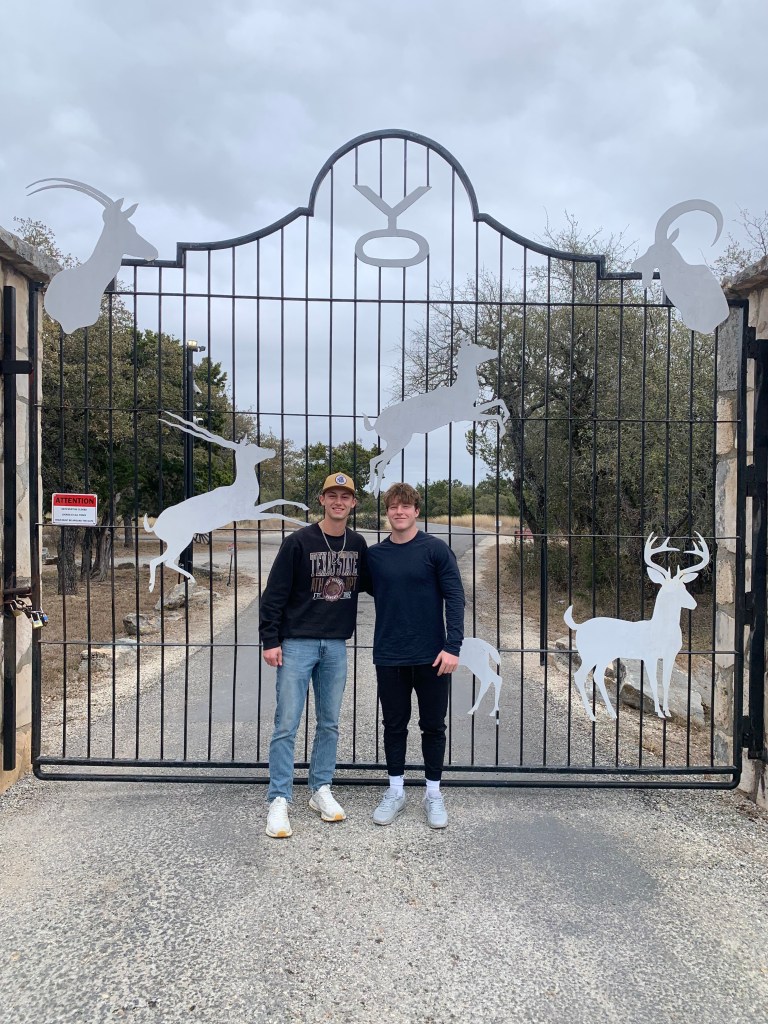 Eric and Kolton are standing in front of the YO metal gate adorned with silhouettes of various animals, including a deer and a goat. Eric and Kolton are posing together, smiling at the camera, dressed in casual attire. 