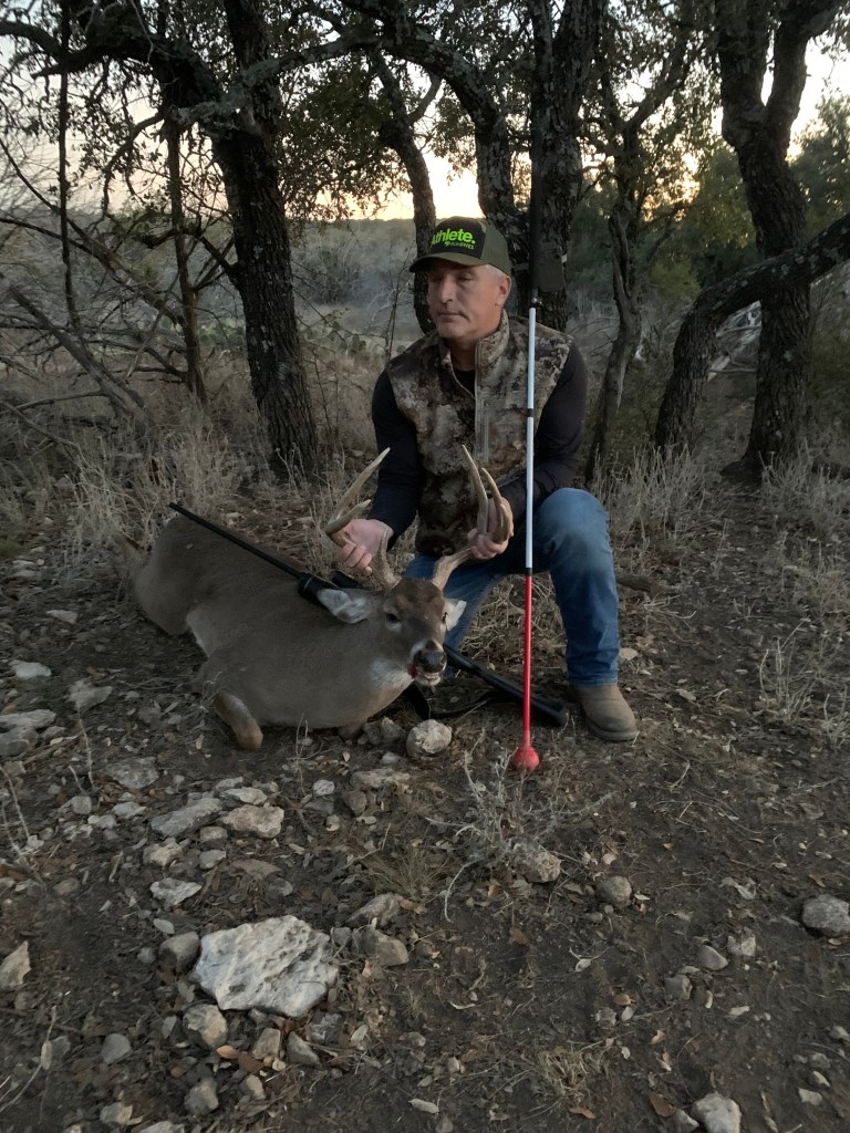 Mike is kneeling in a wooded area, holding up the antlers of a eight-point white tail buck deer lying on the ground. Mike is wearing a camouflage vest, blue jeans, and a camouflage hat.