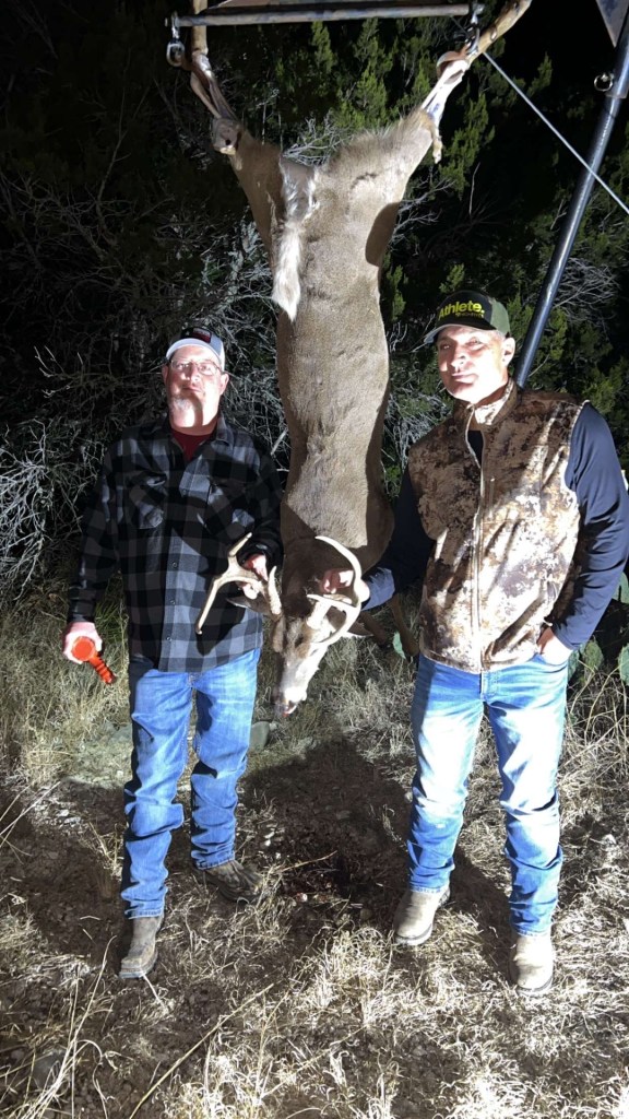 Mike and Kirk are standing next to a eight-point white tail buck deer hanging upside down from a skinner rack. Kirk is wearing a plaid shirt, jeans, and a cap, holding a flashlight in one hand and the deer's antler with the other. Mike is wearing a camouflage vest, long-sleeve shirt, and jeans, also holding the deer. 