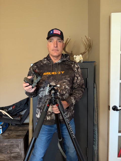 The image shows Mike standing indoors in front of a gun safe. Behind him on top of the safe is a mounted deer skull with antlers. He is wearing a camouflage-patterned jacket with the word "Muddy" printed on it and a Heroes Ranch baseball cap. Mike touches a Bog Deathgrip tripod outfitted with the Barker Outdoors adapter in his left hand and holds the BOG saddle clamp mount in his right.