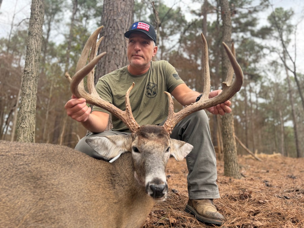 The image shows Mike kneeling in a wooded area, holding the large antlers of a harvested white-tailed deer at Heroes Ranch. The buck deer is referred to as Big 9 by Heroes Ranch hunting guides. Big 9 is lying in front of Mike on the forest floor, which is covered in pine needles.  