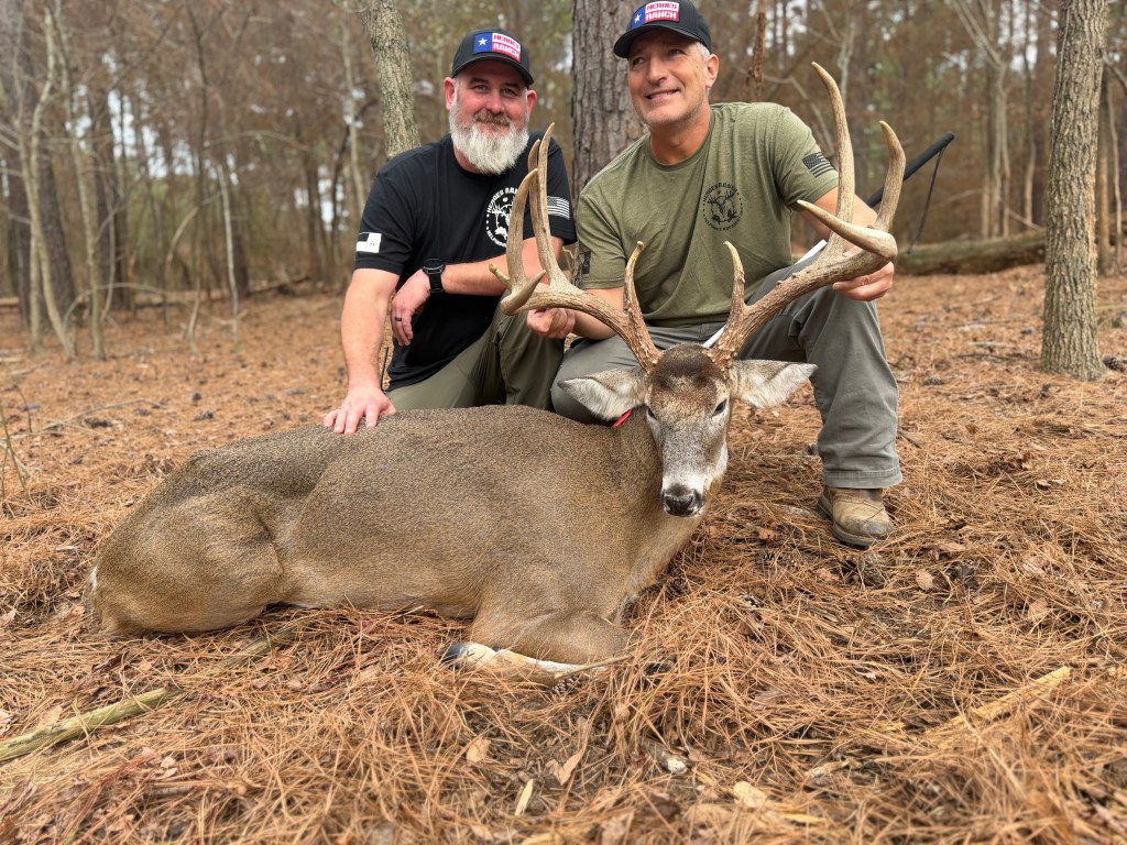 The image shows Luke Baker, Heroes Ranch hunting guide, and Mike. We both wear hats with "Heroes Ranch" written on them and casual outdoor clothing. Between us lies Big 9, a large deer with impressive antlers. Luke has a gray beard and wears a black shirt, while Mike wears a green shirt and beige boots. Mike is holding Big 9’s antlers, and both are smiling at the camera. Trees and natural forest surroundings fill the background.