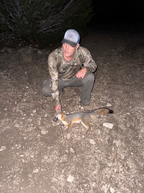 1.	The image shows Mike wearing camouflage clothing and a light blue cap, crouching on a rocky dirt ground. He is holding a small fox. The surrounding area is outdoors, possibly a natural or wilderness setting, and the lighting suggests the photo was taken at night.