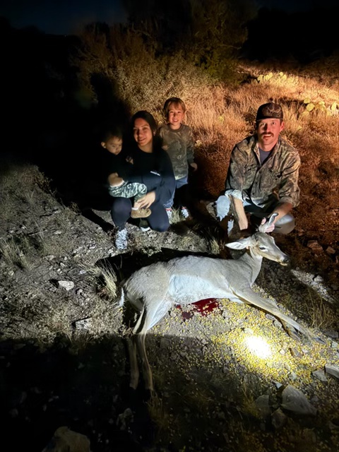 1.	The image shows a nighttime outdoor scene with Kody’s family. They are posing around a whitetail doe deer lying on the ground with a visible wound and some blood. Kody is kneeling and holding the deer's head.