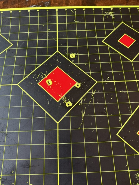 The image shows a black shooting target with yellow grid lines and diamond shapes outlined in yellow. The diamonds have red centers. There are five visible bullet holes grouped around the red center of the main diamond in the middle.