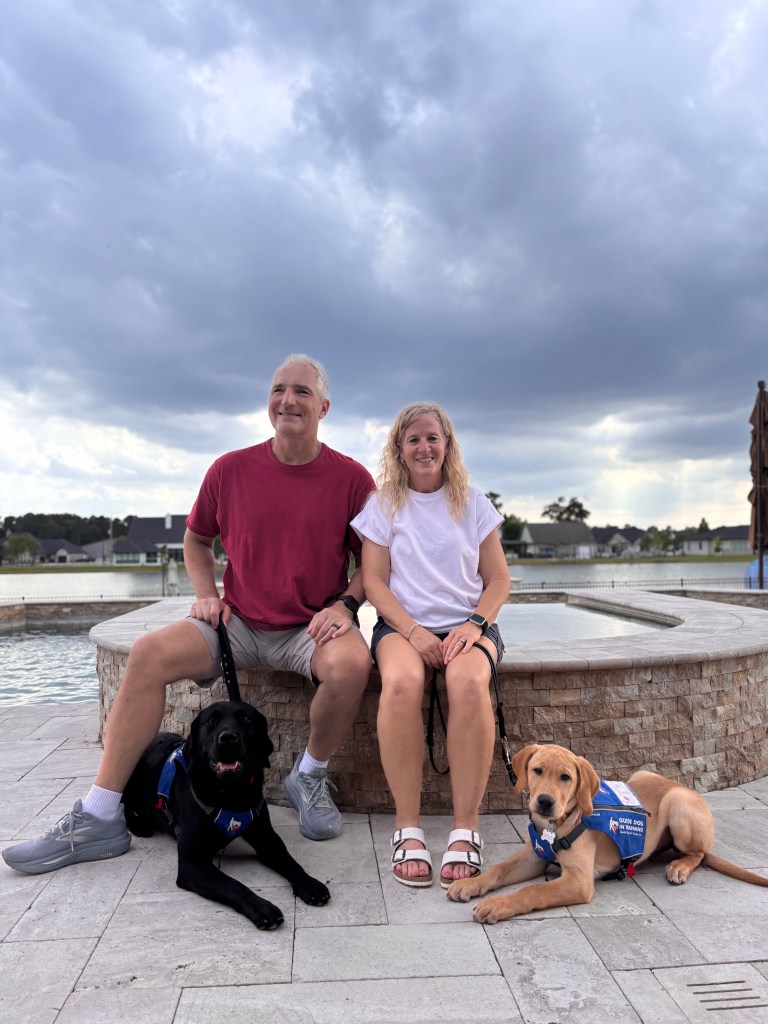 The image shows Mike and Wendy sitting outdoors on the edge of a circular stone water feature. Mike , wearing a red shirt and gray shorts, is seated on the left with Volt lying beside him. Wendy , wearing a white shirt and black shorts, is seated on the right with Xylo lying beside her. Both dogs are wearing blue vests that identify them as guide dogs in training for Guide Dogs of Texas. Volt is the 9th and Xylo is the 10th puppy that Wendy has trained for a guide dog school.