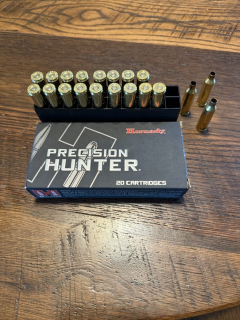 The image shows a box of Hornady Precision Hunter ammunition containing 20 cartridges. The box is dark gray with white and red text. Next to the box, there is a plastic tray holding 17 brass cartridges with visible primers. Additionally, three loose cartridges are standing upright on the wooden surface beside the tray.