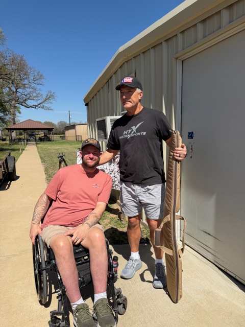 The image shows John Ray and Mike outdoors in front of Sharp Gun Works. We are both wearing Heroes Ranch hats. Mike is standing holding an empty gun case. John Ray is seated in a wheelchair.