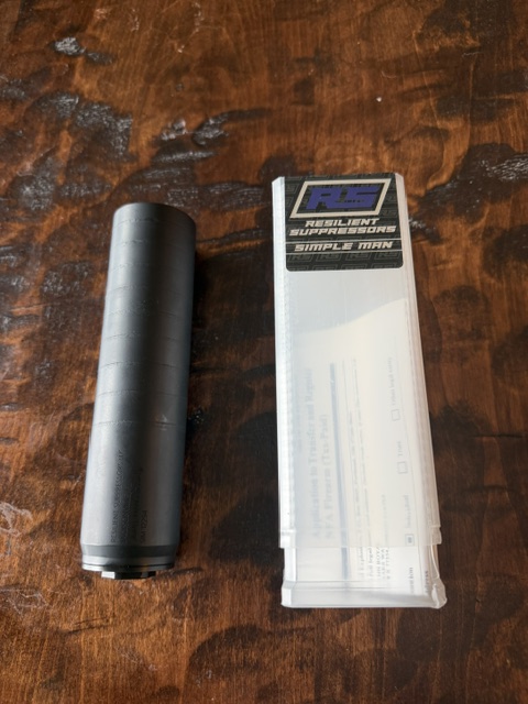 The image shows a Resilient Simple Man suppressor. The black cylindrical object is lying on a wooden surface. Above it is the suppressor’s clear plastic container.