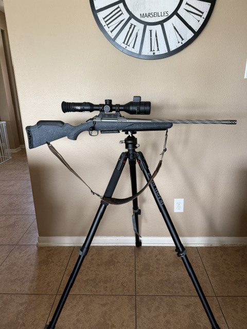 The image shows a black rifle mounted horizontally on a tripod. The rifle has a scope attached.