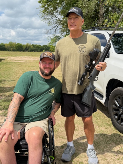 The image shows John Ray and Mike outdoors in a grassy area. Mike is standing on the left wearing a Heroes Ranch T-shirt holding a rifle equipped with a scope. John ray sits in a wheelchair wearing a Heroes Ranch cap.