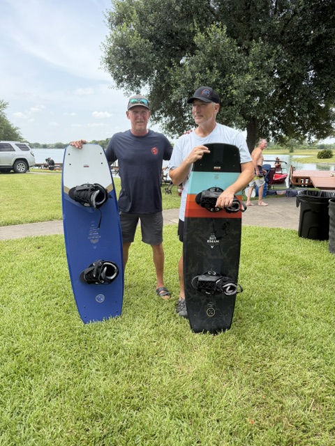 The image shows Russ and Mike standing on grass holding large wakeboards with foot bindings. Russ is on the left wearing a dark shirt, dark shorts, and a visor, holding a blue and white wakeboard. Mike is on the right wearing a white shirt, gray shorts, and a cap, holding a black, teal, and orange wakeboard.