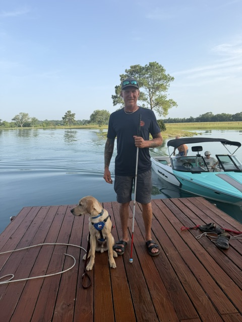 The image shows Russ standing on a wooden dock beside a light brown dog wearing a blue puppy-in-training jacket. Russ is holding a white cane and wearing a black t-shirt, gray shorts, sandals, and a teal cap with sunglasses on top. Behind him is a turquoise boat on the water, and the background features calm water, trees, and a soft blue sky with some clouds. A white coiled rope lies on the dock.