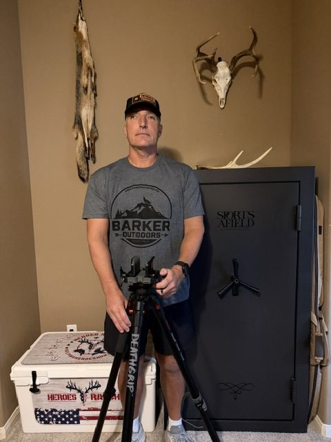The image shows Mike standing indoors in front of a gun safe. Behind him is a mounted deer skull with antlers and a fox skin. He is wearing a Barkers Outdoors shirt and a Heroes Ranch baseball cap.